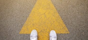 PLM Guide is shown by a man wearing white sneakers standing on a yellow arrow. The picture is taken from a birds eye view only showing his feet on the ground