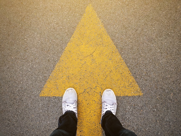 PLM Guide is shown by a man wearing white sneakers standing on a yellow arrow. The picture is taken from a birds eye view only showing his feet on the ground