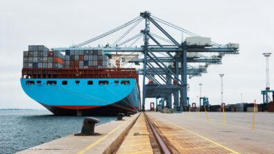 Blue container ship docked at a harbor being serviced by cranes.