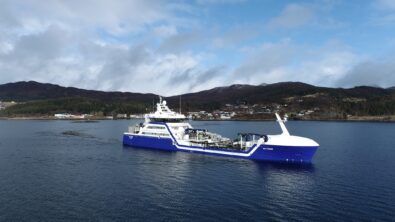 A blue and white ship carrying heavy machinery sails off a coast.