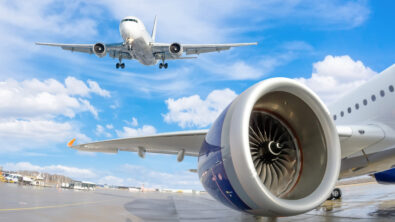 View of an airplane wing and engine with a full airplane flying overhead in the sky.