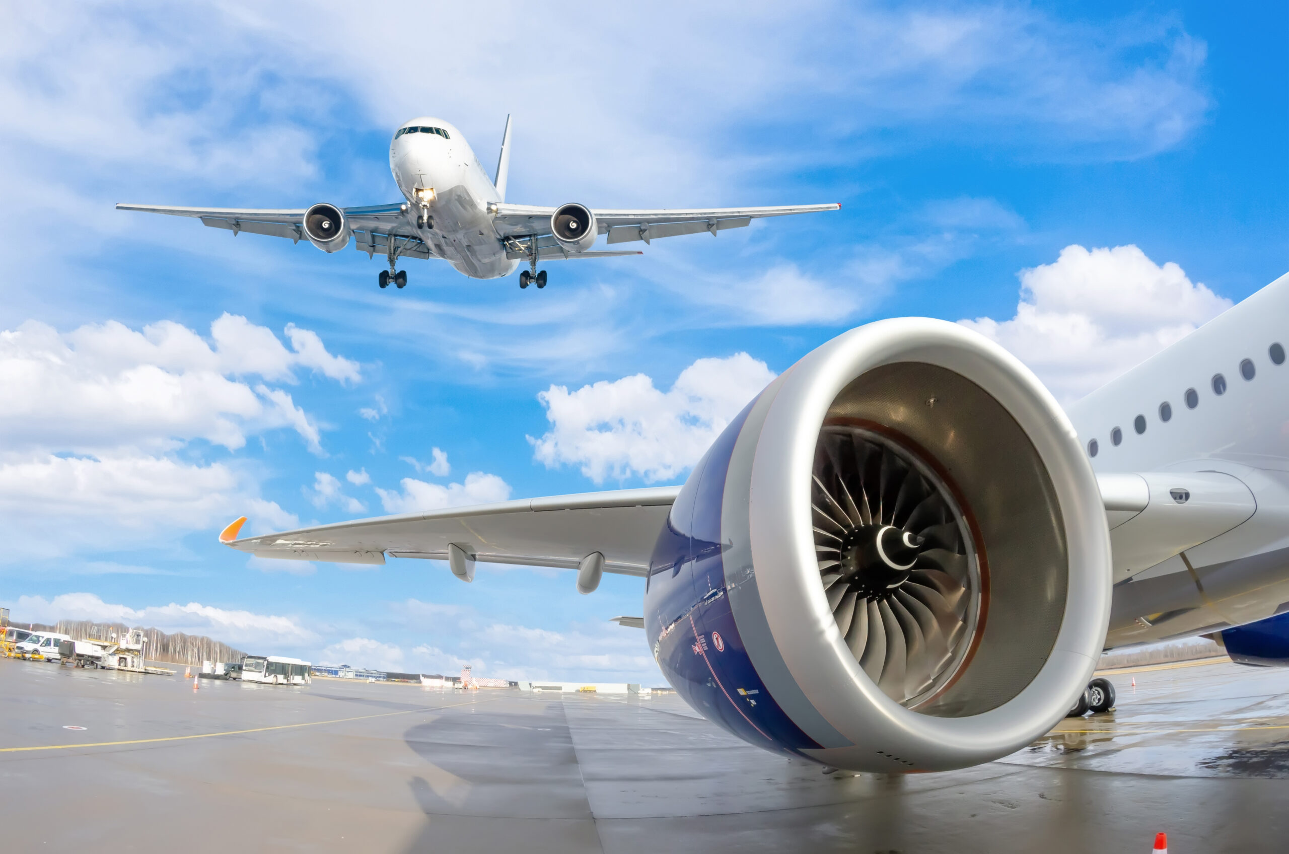 View of an airplane wing and engine with a full airplane flying overhead in the sky.