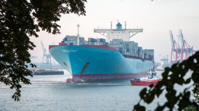 A light blue container ship sailing into a harbor alongside a tug.