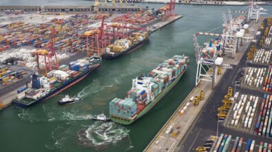 A container ship sailing through a harbor past two other ships.