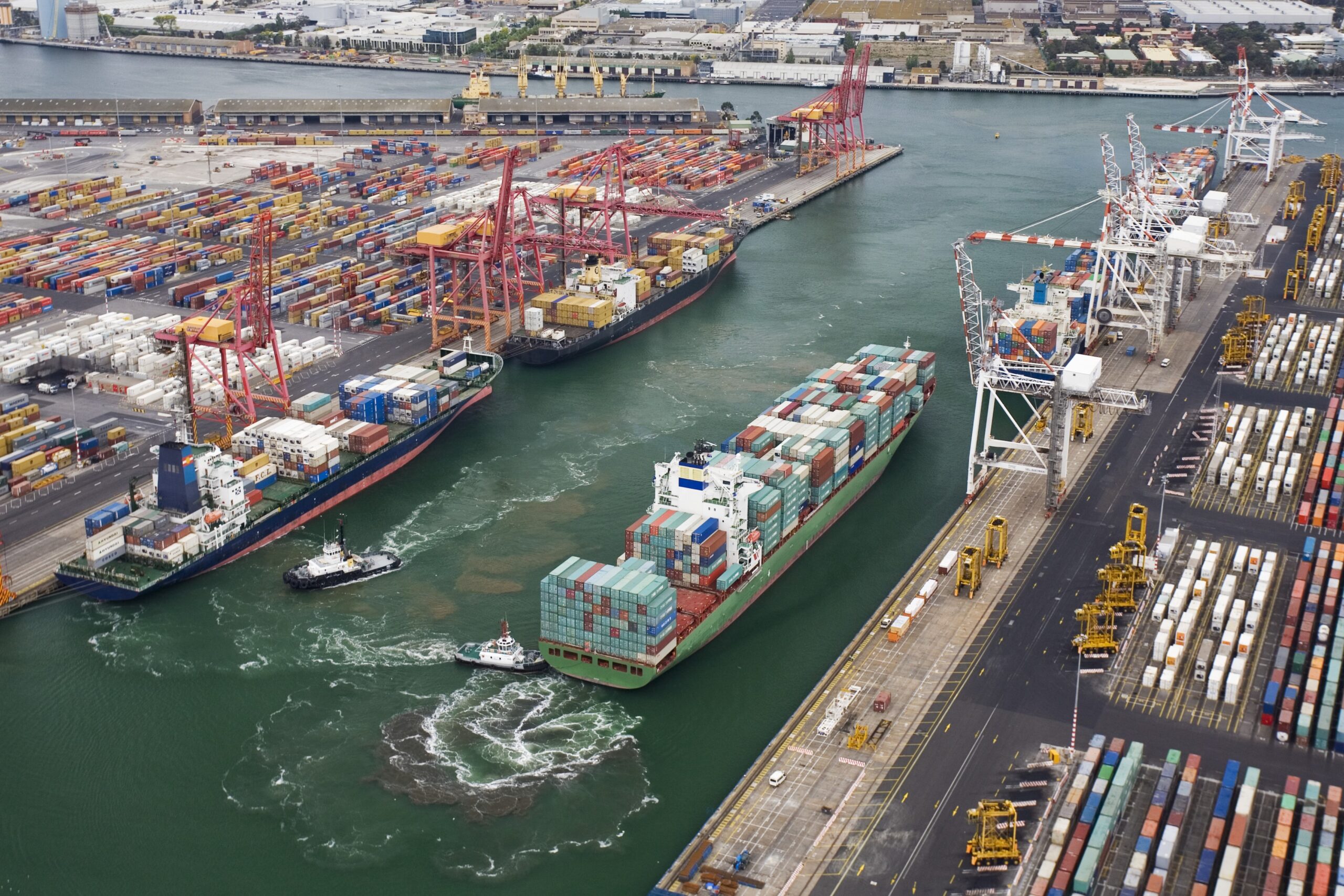 A container ship sailing through a harbor past two other ships.