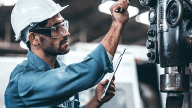 A factory worker consults a tablet computer while working on a machine. The ability for apps built using low-code platforms enables servitization in the manufacturing vertical.