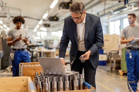 A man wearing a suit in a factory looks at laptop screen, highlighting the need for customized PLM solutions.