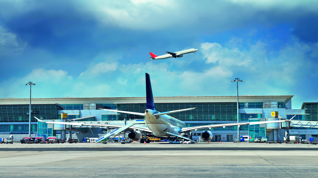 An airplane sits at the gate at the airport as another plane flies through the air in the background