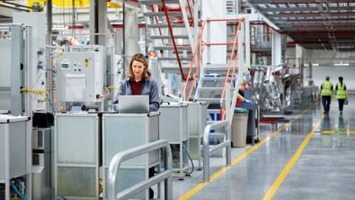 Woman at a standing computer terminal in the middle of a manufacturing plant