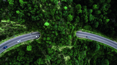 A bird's eye view of a road curving through a green forest, with a few cars driving along it.