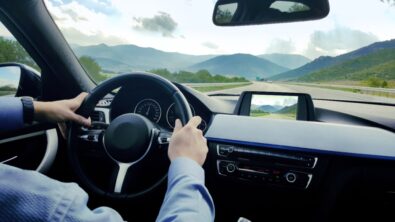 A car dashboard with a person behind the steering wheel, driving on a road through a hilly landscape.