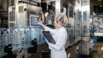 A worker in a white lab coat and hairnet operates a touchscreen interface on a large, industrial bottling machine.