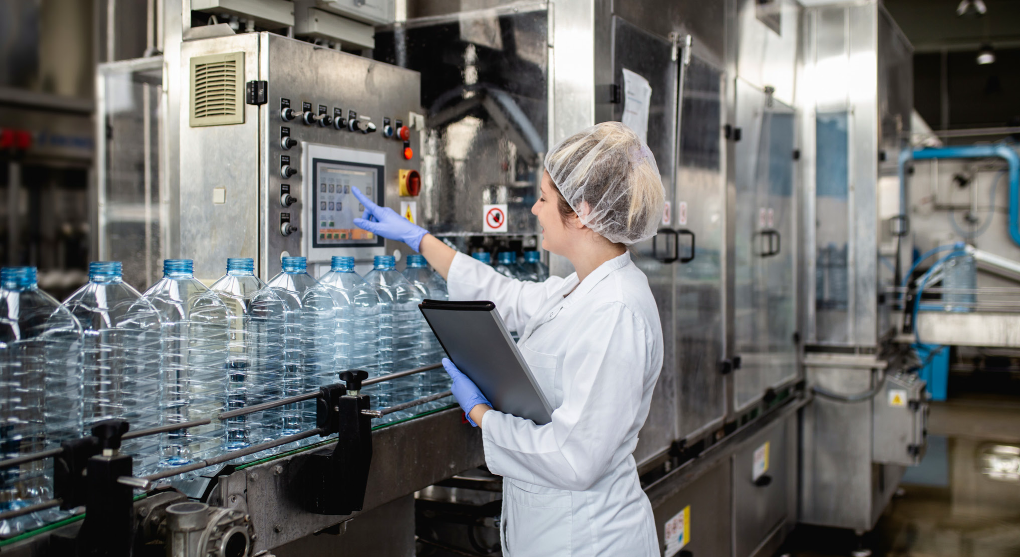 A worker in a white lab coat and hairnet operates a touchscreen interface on a large, industrial bottling machine.