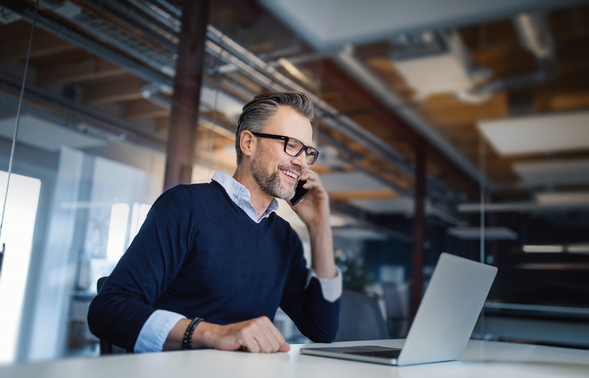 Mid adult business professional sitting at table with laptop and talking on mobile phone. Businessman working in office.