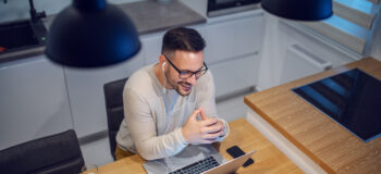 Top view of attractive smiling caucasian man sitting at dining table in kitchen and having video call with his girlfriend. On table are laptop, glass with water and mug with coffee.