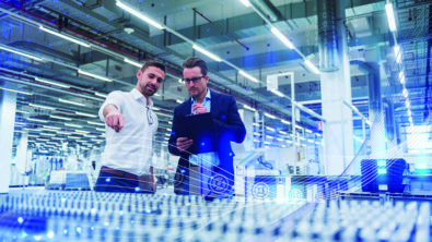 Two business men standing in a factory. One is pointing and the other takes notes on a tablet.