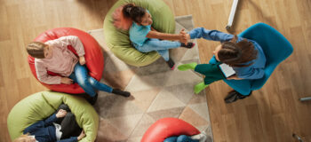 Top View Shot in Elementary School Creativity Classroom: Children Sitting on the Bean bags while Teacher Explains Lesson to them and Uses Digital Tablet Computer, They Play by Passing High Five.